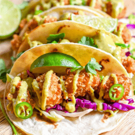 Three easy fried chicken tacos filled with crispy fried toppings, shredded cabbage, sliced jalapeños, cilantro, and drizzled sauce, garnished with lime wedges.