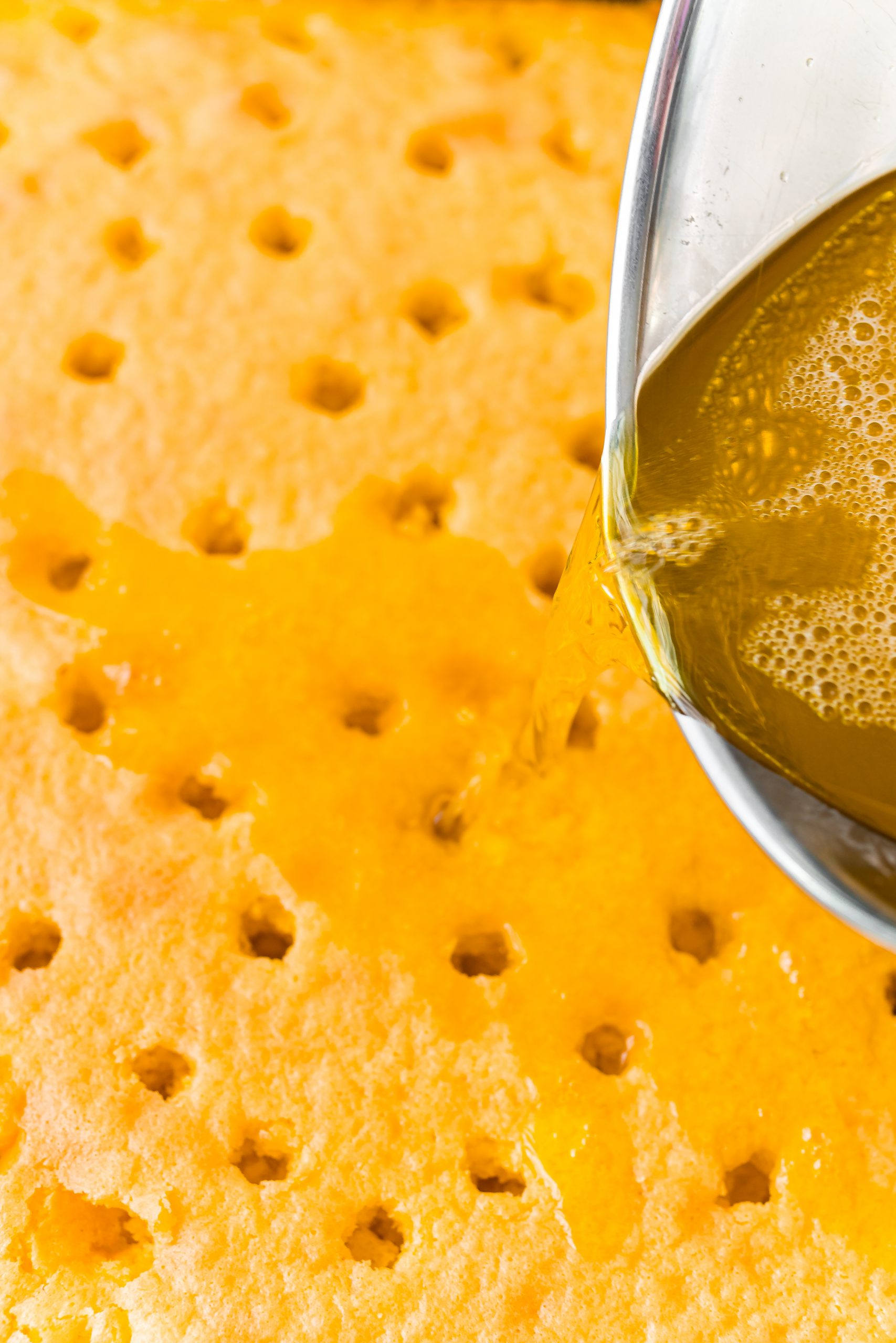 A spoon is being used to pour lemon Jello onto a Lemon Poke Cake.