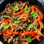 A skillet filled with cooked beef strips, red bell peppers, onions, and sliced green jalapenos creates the perfect one pot steak fajitas.