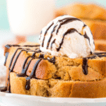 A slice of peanut butter dessert bread topped with a scoop of vanilla ice cream and drizzled with chocolate syrup on a white plate.