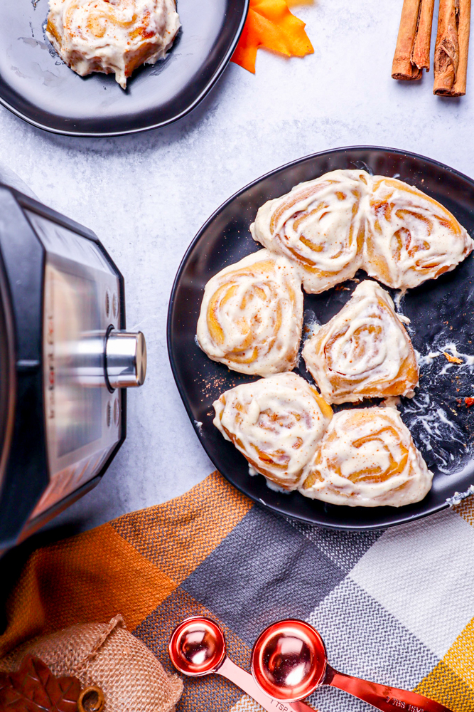 pumpkin cinnamon rolls in a black pan sitting next to a black and silver instant pot on a marble table with a checkered hand towel