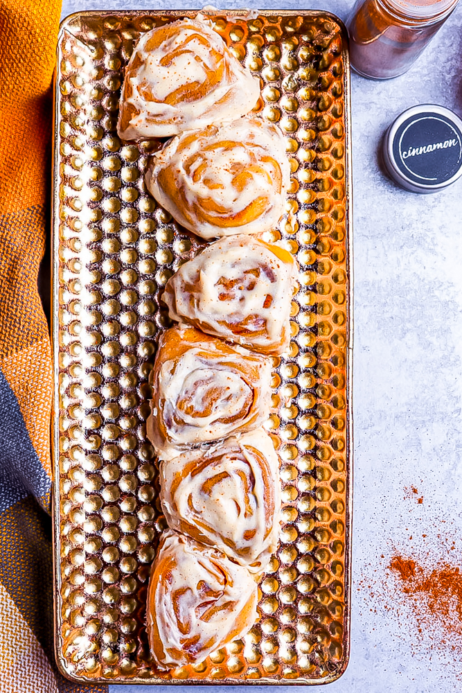 pumpkin cinnamon rolls sitting longways on a golden plate that has lots of perforations. Cinnamon in the background on a marble table 