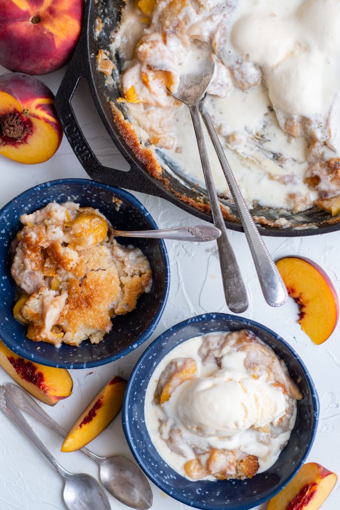 picture of peach cobbler and ice cream in a skillet and bowls with spoons and peaches on the table