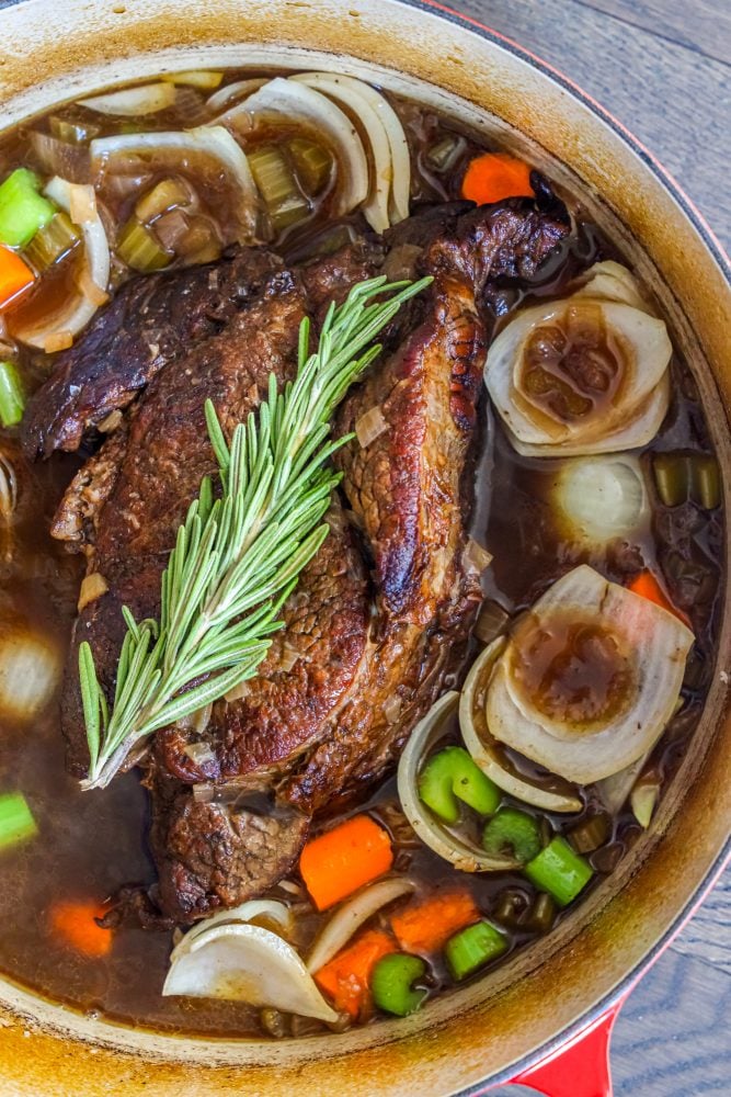 pot roast in dutch oven with gravy and carrots, celery, onions, and rosemary on top 