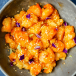 A bowl of breaded shrimp coated in an orange glaze, garnished with sesame seeds and small purple cabbage pieces, inspired by The Best Easy Bang Bang Shrimp Recipe - Bonefish Grill Copycat.