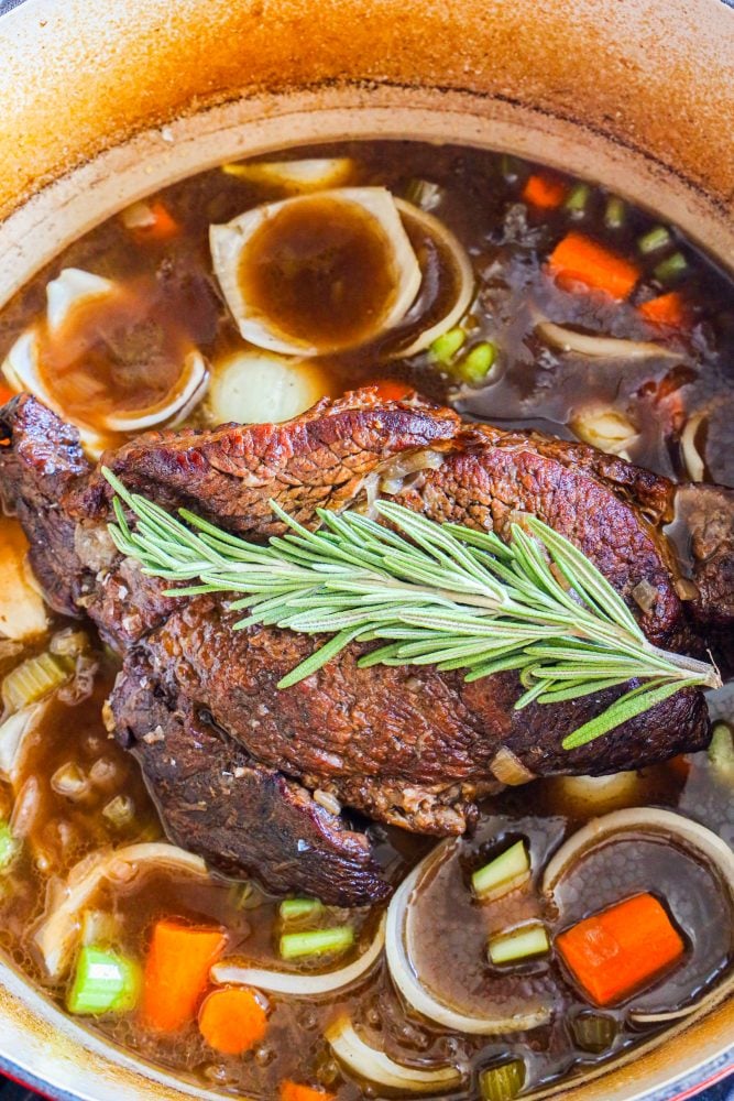 pot roast in dutch oven with gravy and carrots, celery, onions, and rosemary on top 