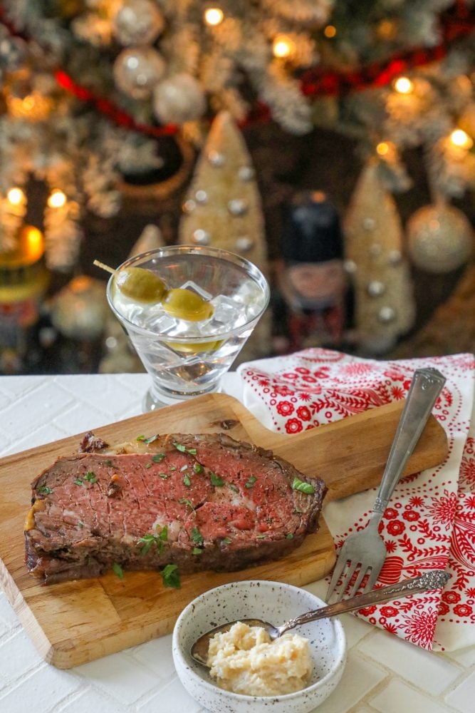 roasted prime rib sliced on a wood cutting board next to a Christmas tea towel and a martini with ice and olives in it