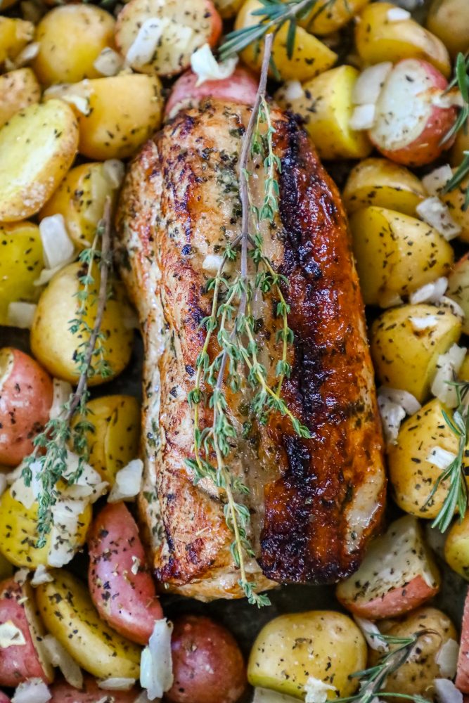 picture of baked pork loin in a pan with seasoned potatoes and onions and a sprig of rosemary