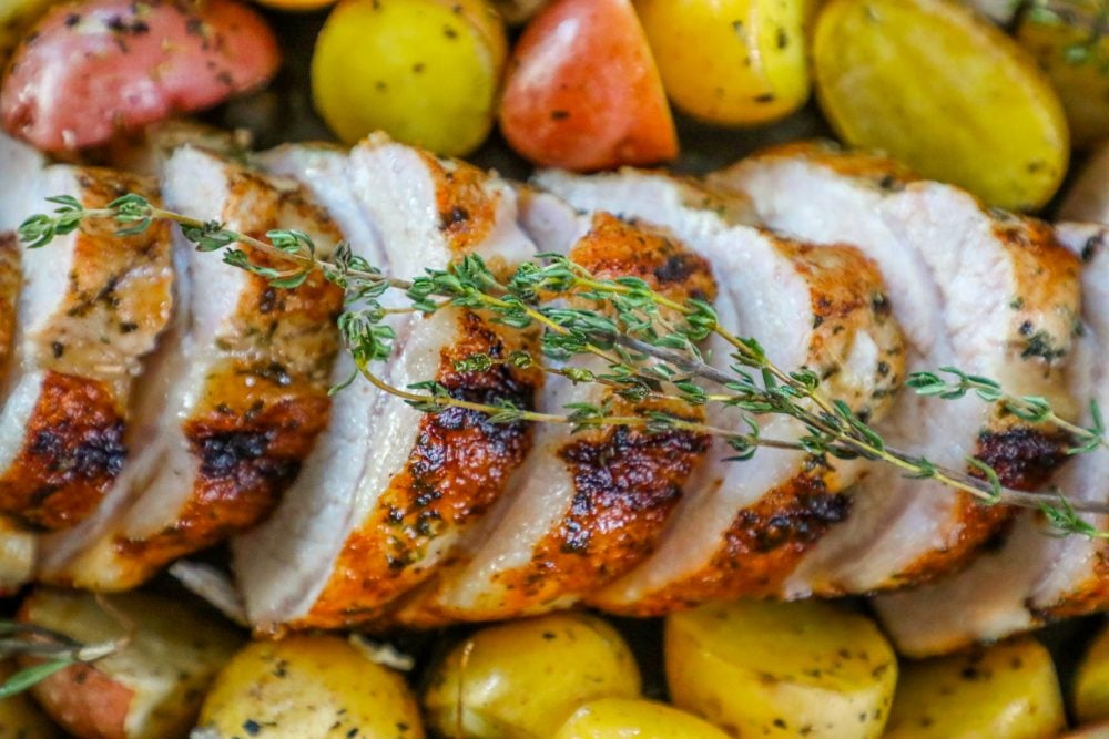 picture of baked pork loin in a pan with seasoned potatoes and onions and a sprig of rosemary