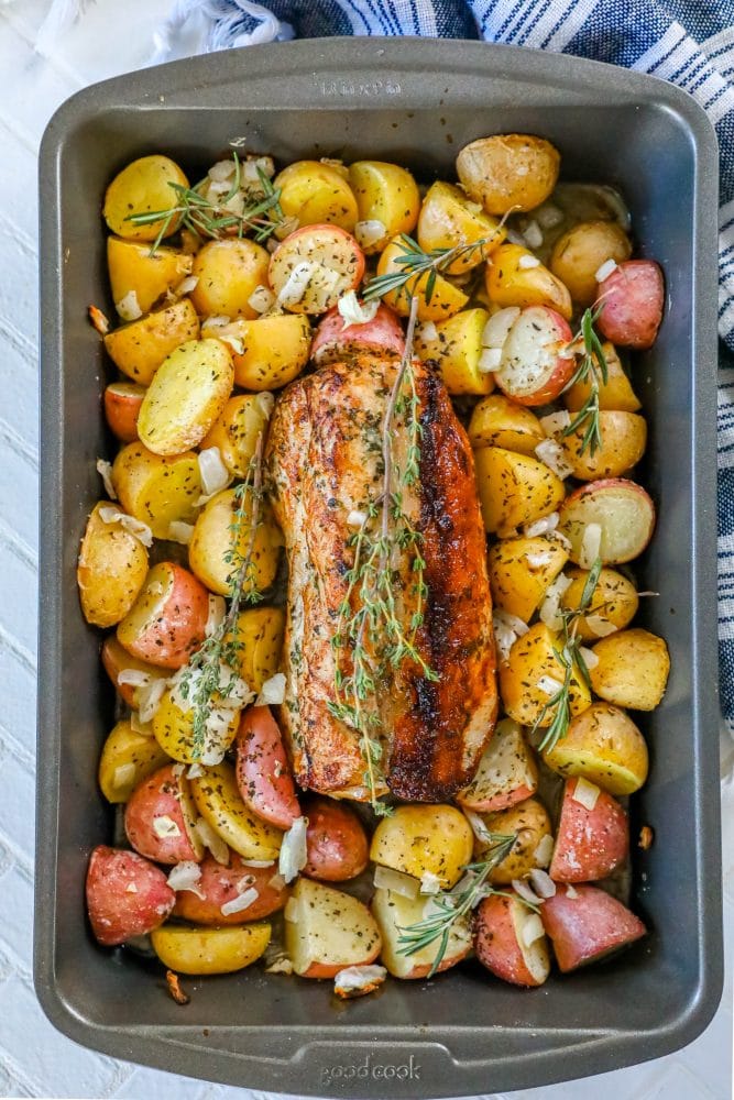 picture of baked pork loin in a pan with seasoned potatoes and onions and a sprig of rosemary