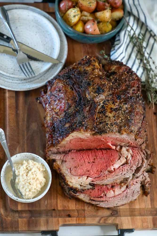 roasted prime rib sliced on a wood cutting board next to horseradish and a bowl of roasted potatoes