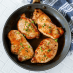 Three garlic butter pork chops garnished with herbs in a skillet, on a white and blue striped towel.