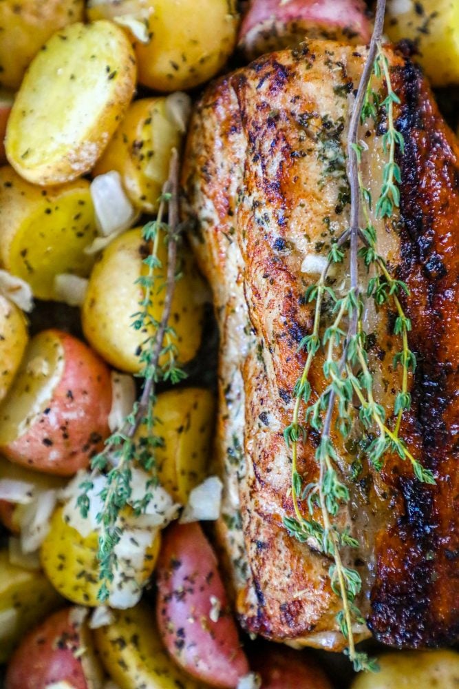 picture of baked pork loin in a pan with seasoned potatoes and onions and a sprig of rosemary