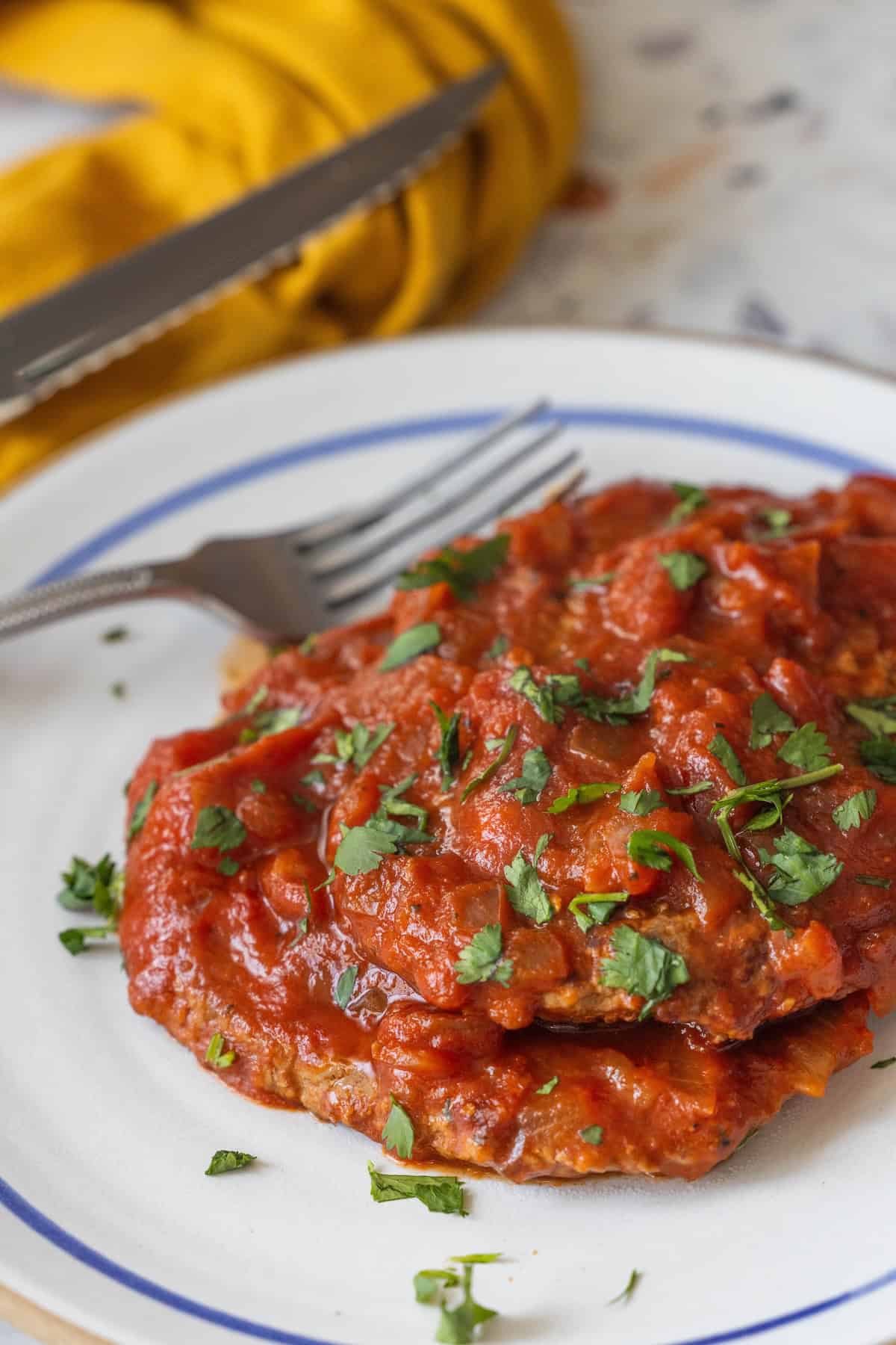 A plate with two pieces of meatloaf, reminiscent of a Swiss Steak, topped with tomato sauce and chopped herbs, next to a fork.