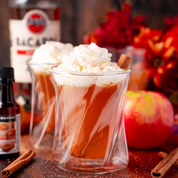 Two glasses of hot apple cider with whipped cream and cinnamon sticks.