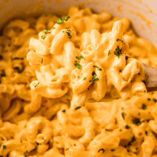 Close-up of creamy macaroni and cheese with sprinkled parsley, shown in a pot with a wooden spoon scooping it. This easy macaroni and cheese recipe incorporates rich pub cheese for an extra indulgent twist.