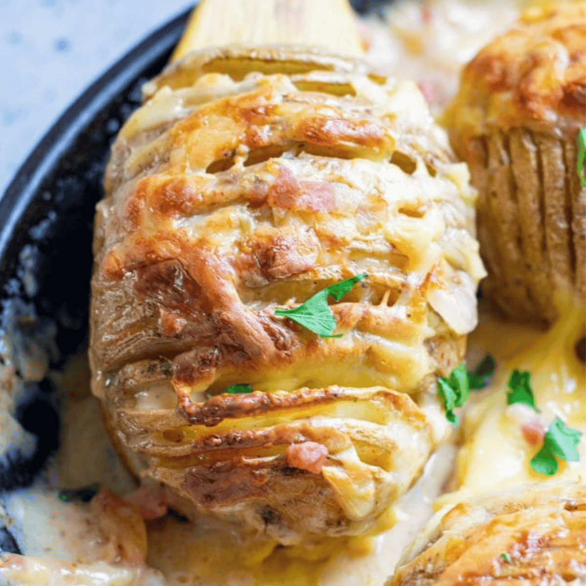 Close-up of creamy cheddar and bacon hasselback potatoes topped with melted cheese and sprinkled with parsley, nestled in a skillet with a luxurious sauce.