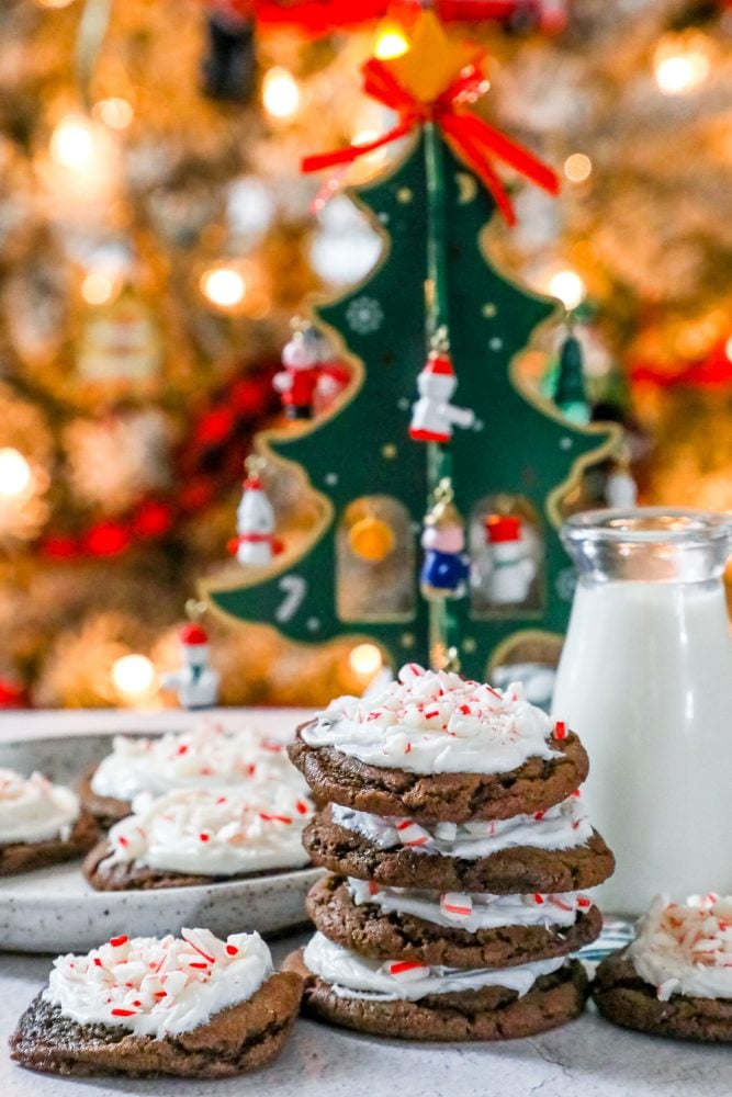 picture of chocolate cookies with frosting and pieces of crushed peppermint candy canes milk and Christmas trees in the background.