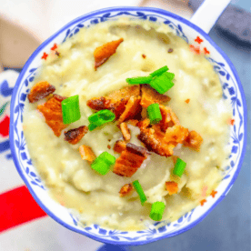 A bowl of creamy, slow-cooker leftover mashed potato soup topped with crispy bacon pieces and chopped green onions, served in a decorative blue and white bowl.