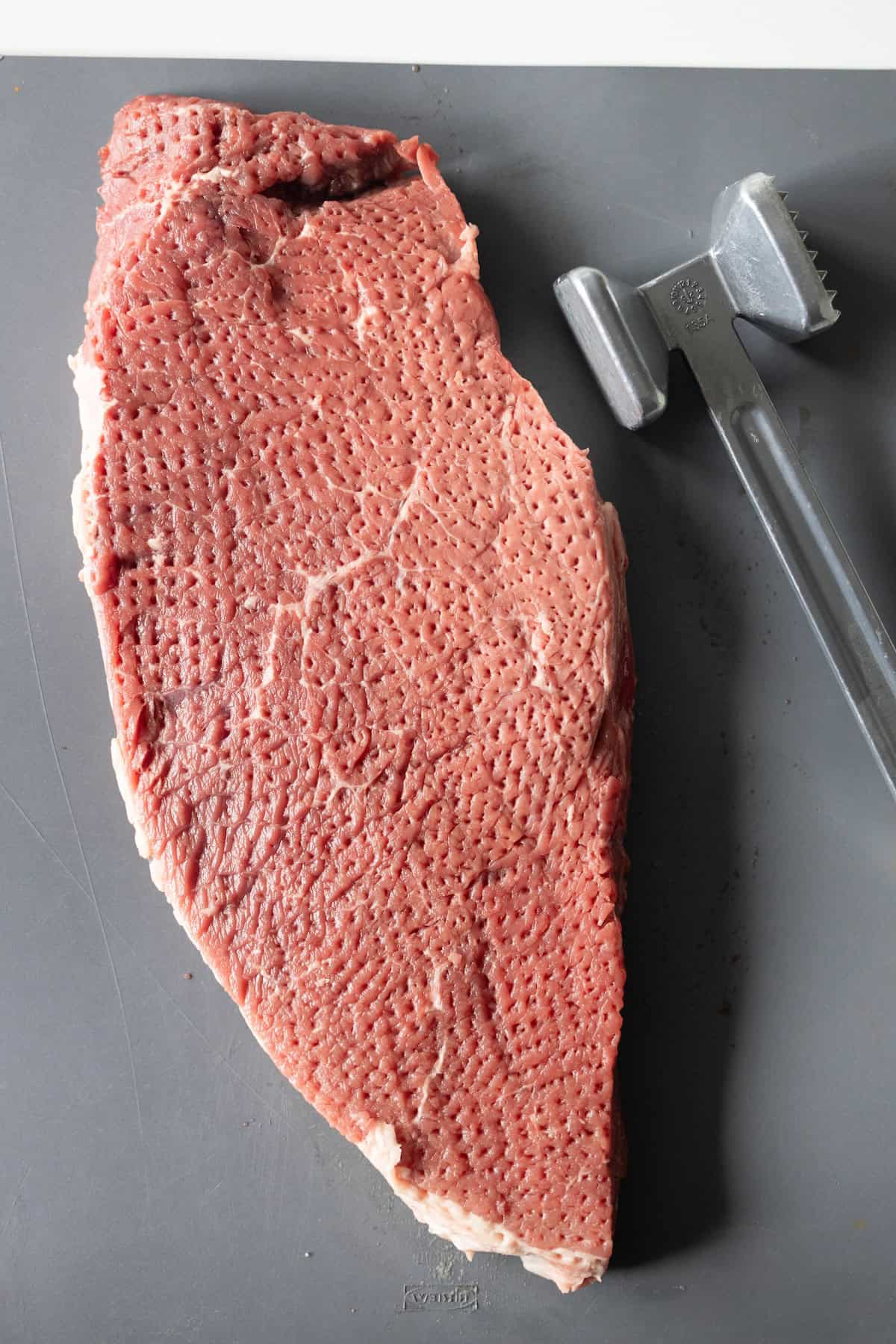A raw broiled top round steak with a textured surface sits on a gray cutting board next to a metal meat tenderizer, perfect for your best easy London broil.