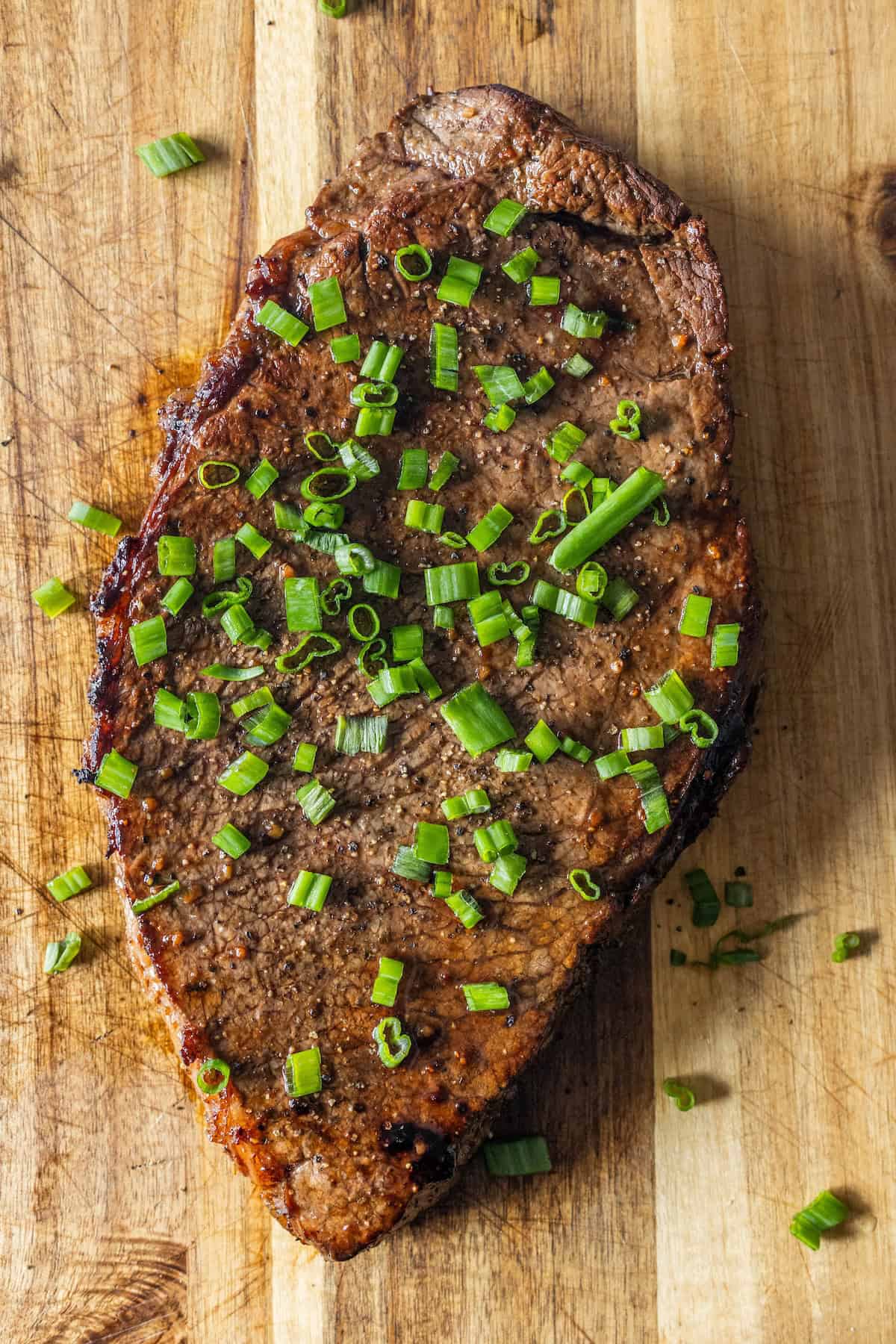 A cooked Keto London broil steak garnished with chopped green onions sits on a wooden cutting board.