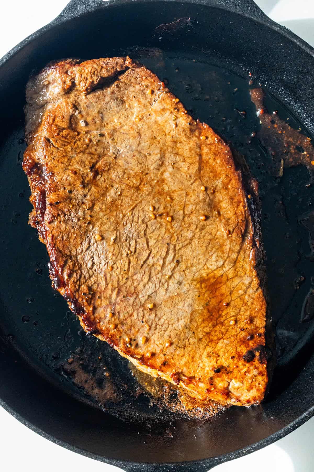 A cooked steak sits in a black cast iron skillet with visible seared marks and some oil around the edges, showcasing an easy London broil that's perfect for savoring at home.