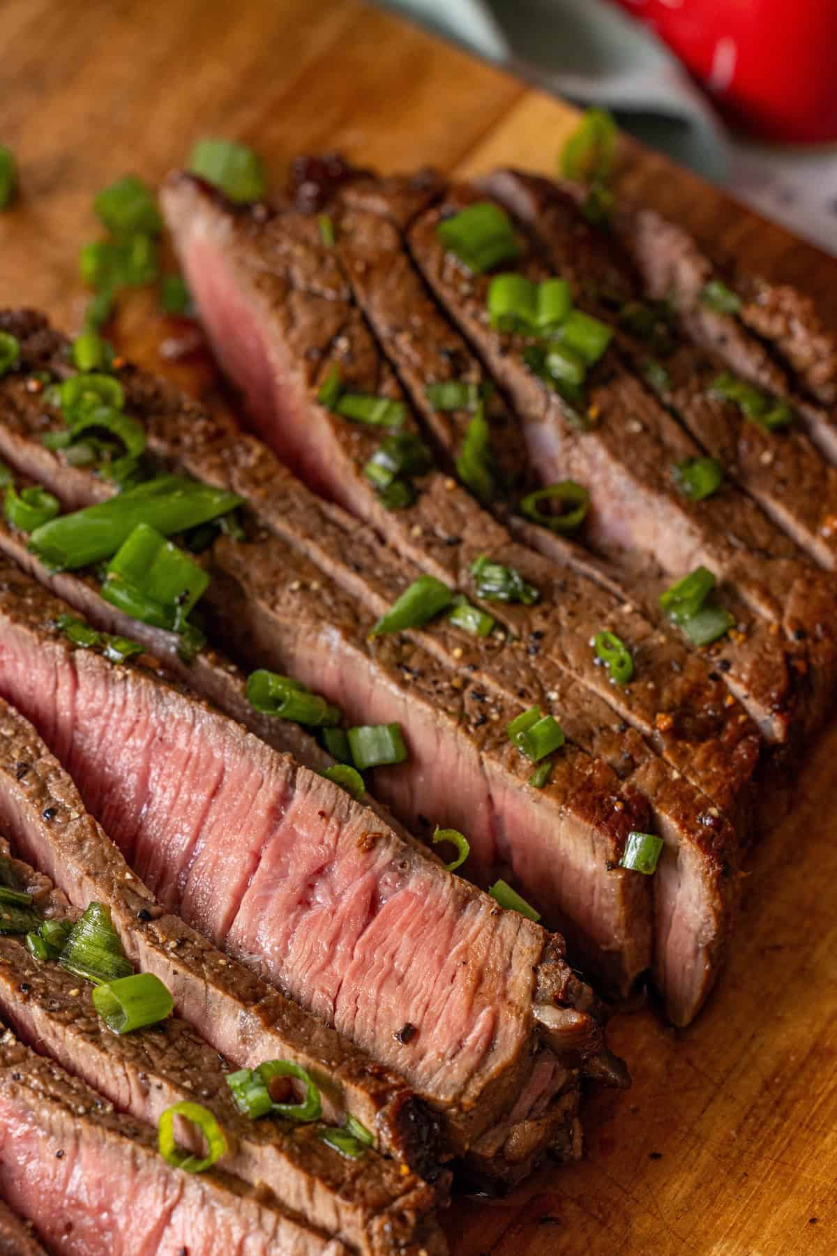 Sliced medium-rare Keto London broil steak topped with chopped green onions on a wooden cutting board.