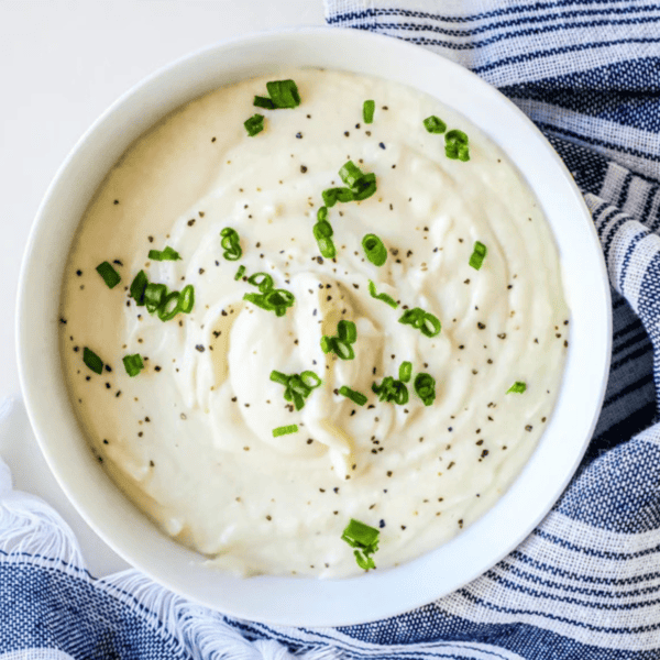 A bowl of whipped cauliflower, inspired by The Best Easy Keto Mashed Cauliflower Recipe, garnished with chopped green onions and black pepper, placed on a blue and white striped cloth.
