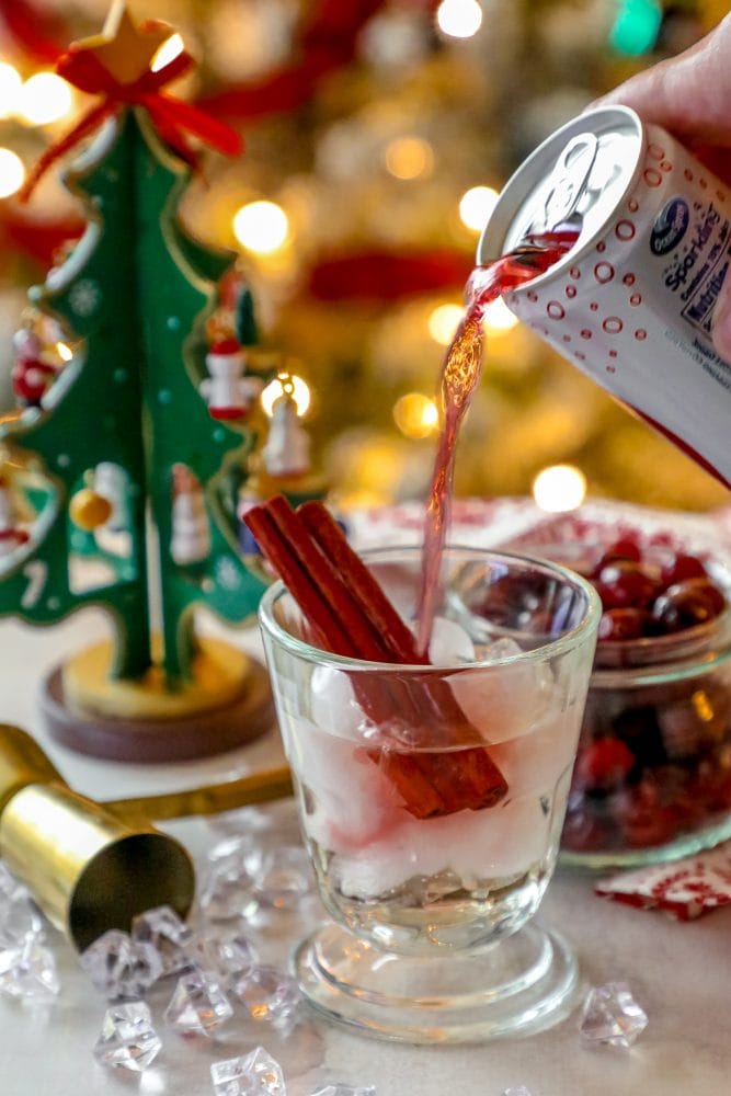 glass of cranberry juice being poured into a glass with cinnamon sticks, ice, and cranberries in it