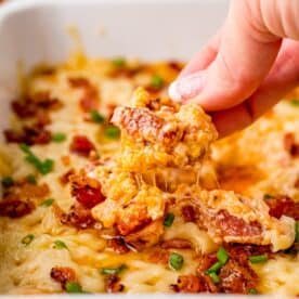 A person preparing a savory bacon and cheese dip by dipping a piece of bacon and cheese casserole into a dish.