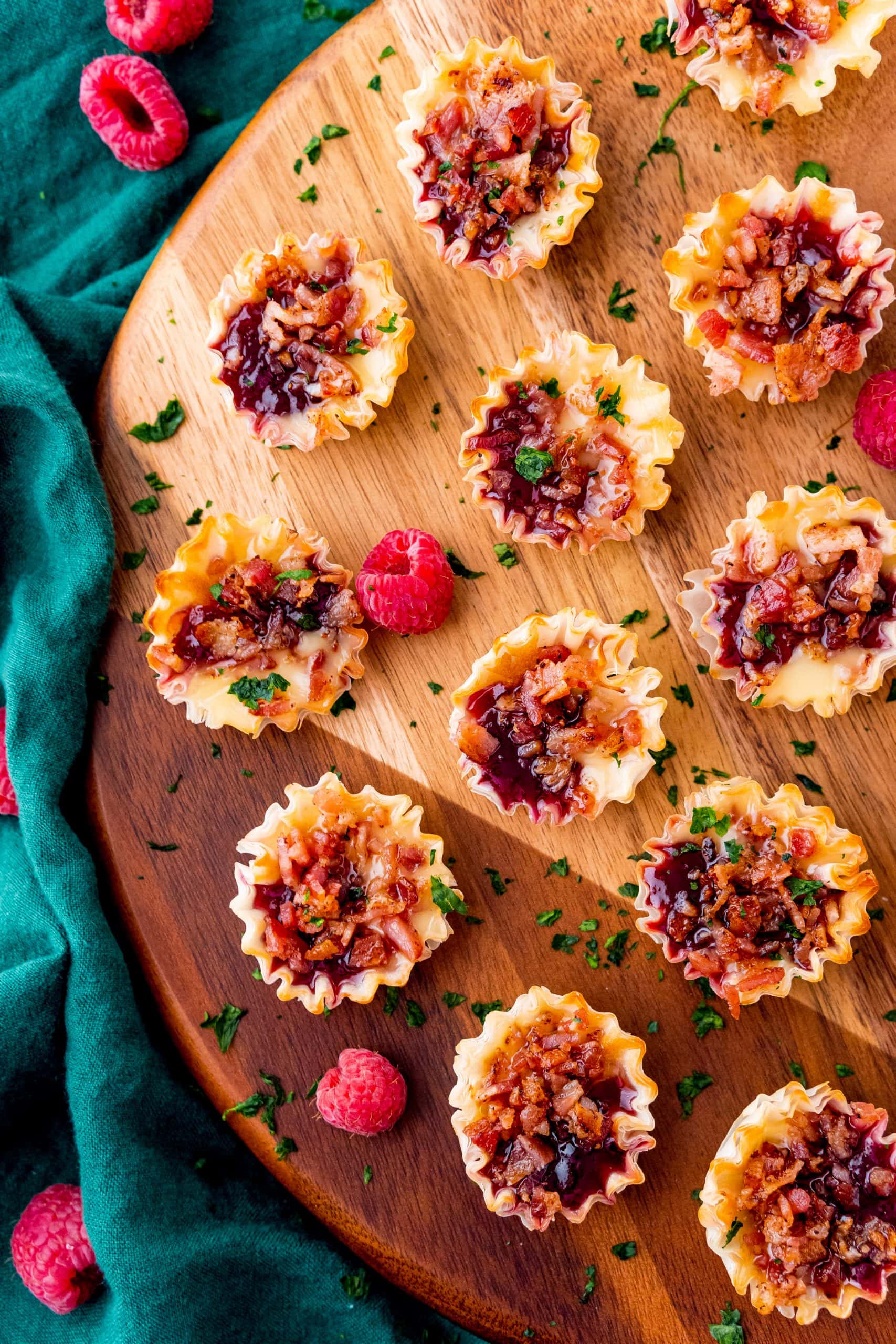 Close-up of golden phyllo cups with melted cheese, ruby-red raspberry jam, and savory bacon pieces on a wooden board.