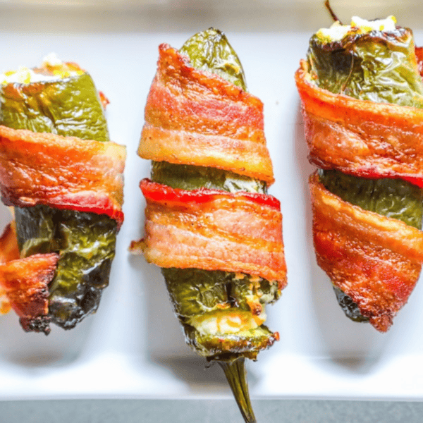 Three bacon-wrapped cheddar and garlic jalapeño poppers filled with cheese sit on a white plate, offering an easy low-carb treat perfect for keto enthusiasts.