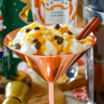 A copper martini glass brimming with whipped cream, caramel sauce, coffee beans, and cinnamon sticks evokes the delicious elegance of a Caramel Espresso Martini. In the blurred background, a bottle and festive Christmas decorations add to the seasonal allure.