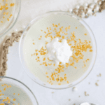A close-up of a dessert in a clear glass cup, topped with a dollop of whipped cream and sprinkled with golden sugar crystals, reminiscent of champagne jello shots, placed on a white surface with a textured fabric edge.