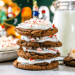 Stack of iced peppermint chocolate cookies topped with crushed candy canes on a counter next to a glass of milk and a bowl of more cookies.