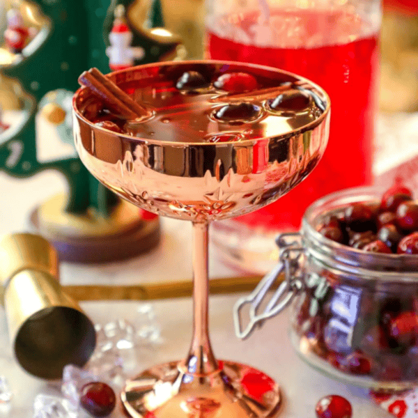 A rose gold cocktail glass filled with a red drink, cranberries, and a cinnamon stick, surrounded by cranberries, a jar, and festive decorationsโperfect for serving an easy cranberry sangria at your next holiday gathering. This elegant scene beautifully showcases the classic Christmas sangria recipe in style.