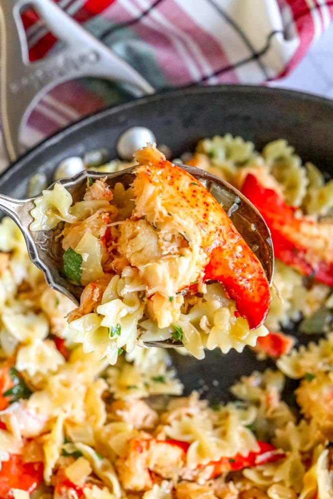 a spoon scooping lobster meat pasta over a pan of bowtie pasta with herbs, cheese, and lobster meat