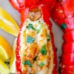lobster tails seasoned with paprika and butter on a white plate with chopped parsley on top with lemons on the plate next to it