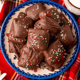 A plate of chocolate-covered square treats, reminiscent of easy millionaires bars, topped with sprinkles and sea salt sits on a red plaid tablecloth.
