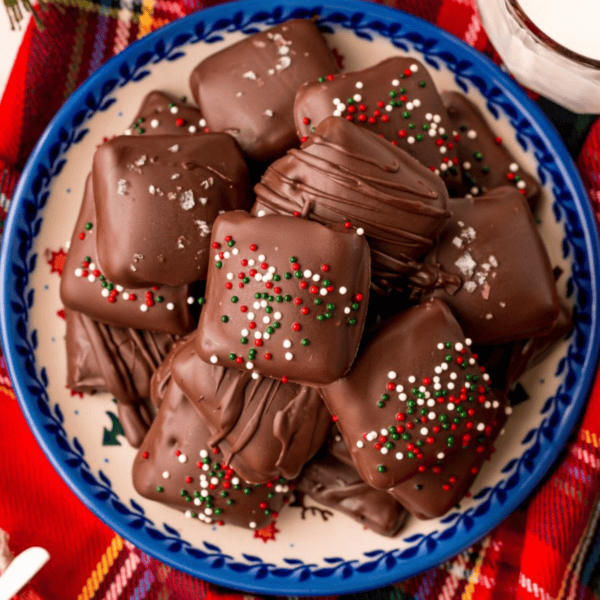 A plate of chocolate-covered square treats, reminiscent of easy millionaires bars, topped with sprinkles and sea salt sits on a red plaid tablecloth.