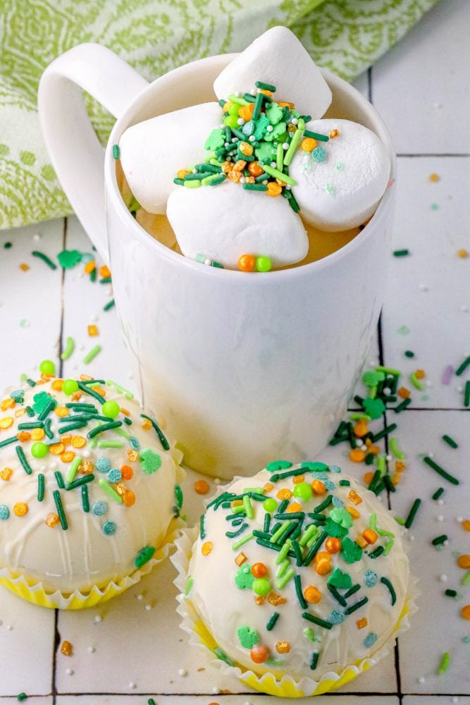 white chocolate hot chocolate bombs covered in saint patricks day sprinkles on a table next to a mug with marshmallows and green and gold sprinkles in it