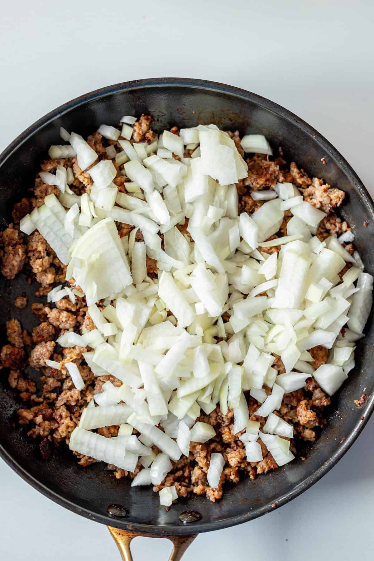 Chopped onions are placed on top of cooked ground meat in a black skillet, ready to complement a ravioli with tomato spinach cream sauce for a flavorful meal.
