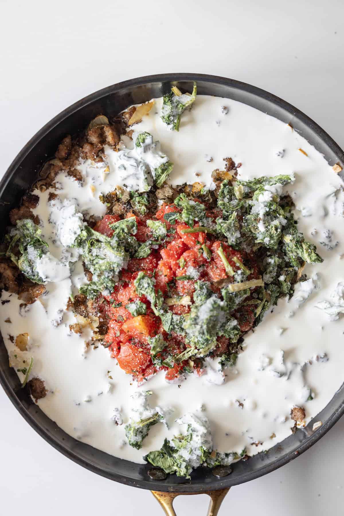 A skillet filled with cooked ground meat, chopped spinach, diced tomatoes, and cream creates the base for a delicious spinach tomato cream sauce recipe, ready to be mixed and enjoyed.