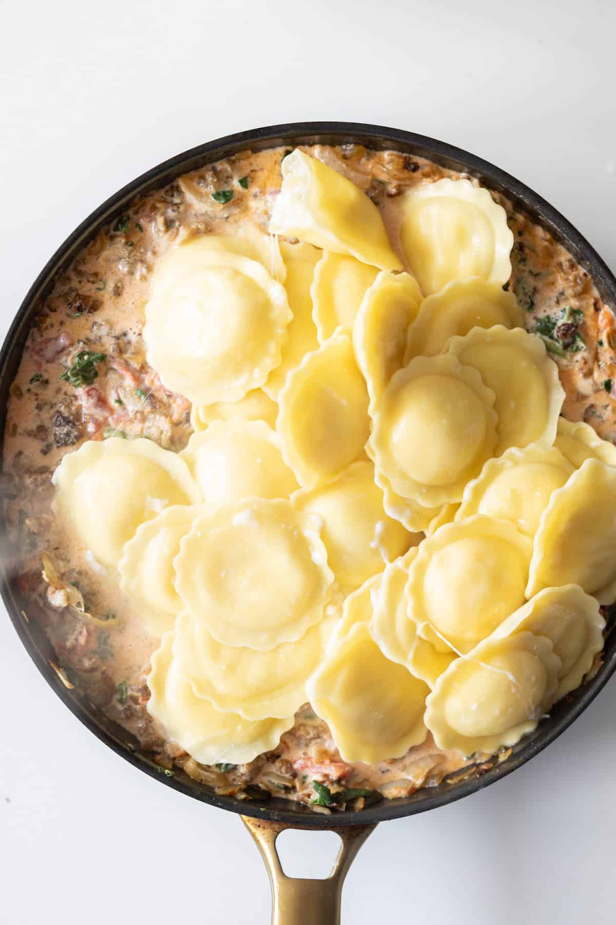A skillet filled with stuffed ravioli sits atop a creamy spinach tomato cream sauce with herbs and diced vegetables.