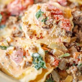 Close-up of ravioli with tomato spinach cream sauce, ground meat, fresh spinach, tomatoes, onions, and a creamy base, topped with grated cheese and red pepper flakes.
