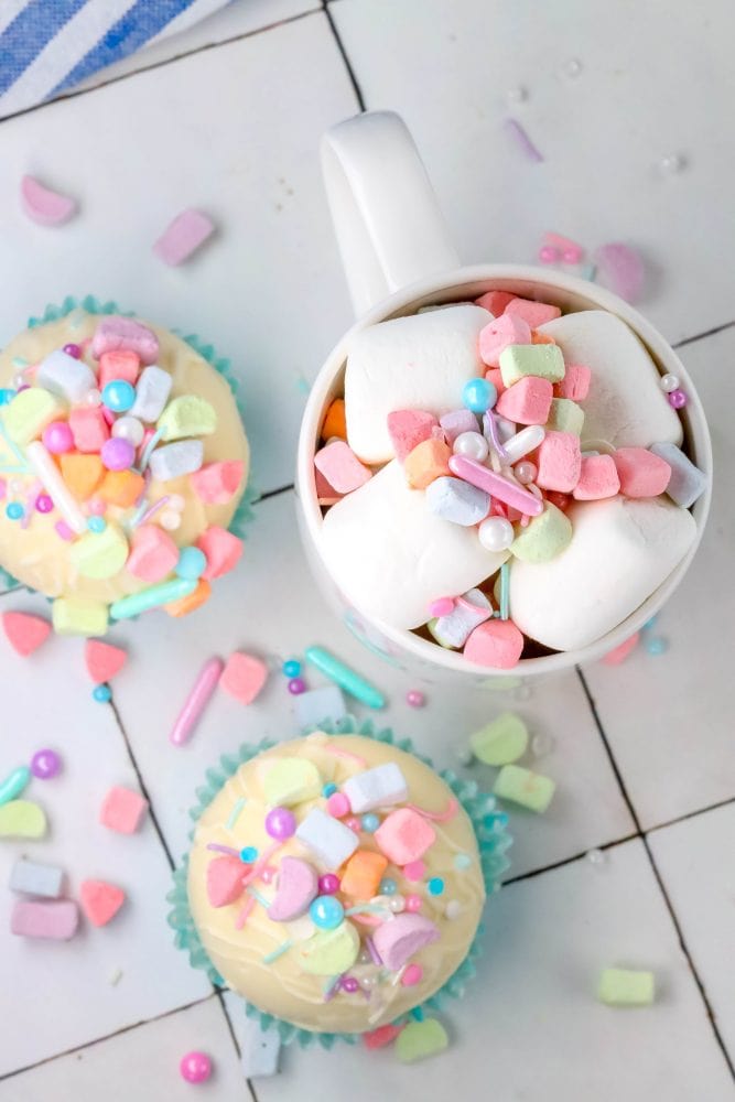 white chocolate hot chocolate bomb in a blue cupcake wrapper with lucky charms marshmallows and sprinkles on top next to a mug with cocoa, sprinkles, and marshmallows in it