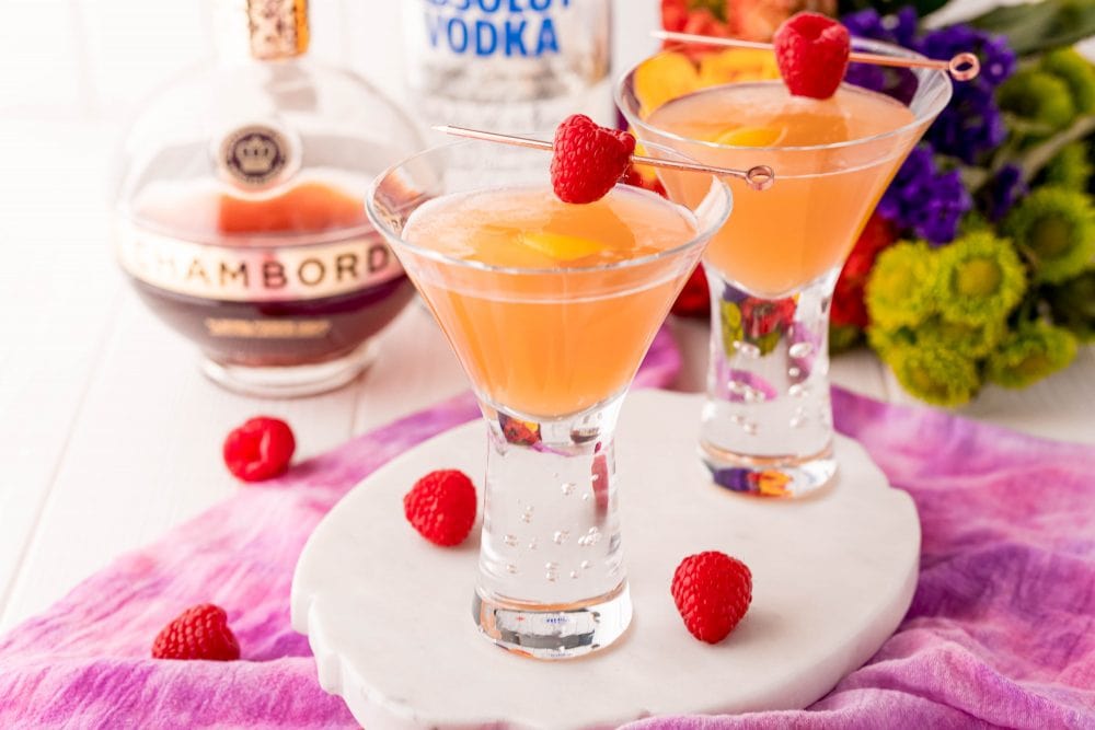 orange martini in a glass with a raspberry stuck across it and chambord in the background.