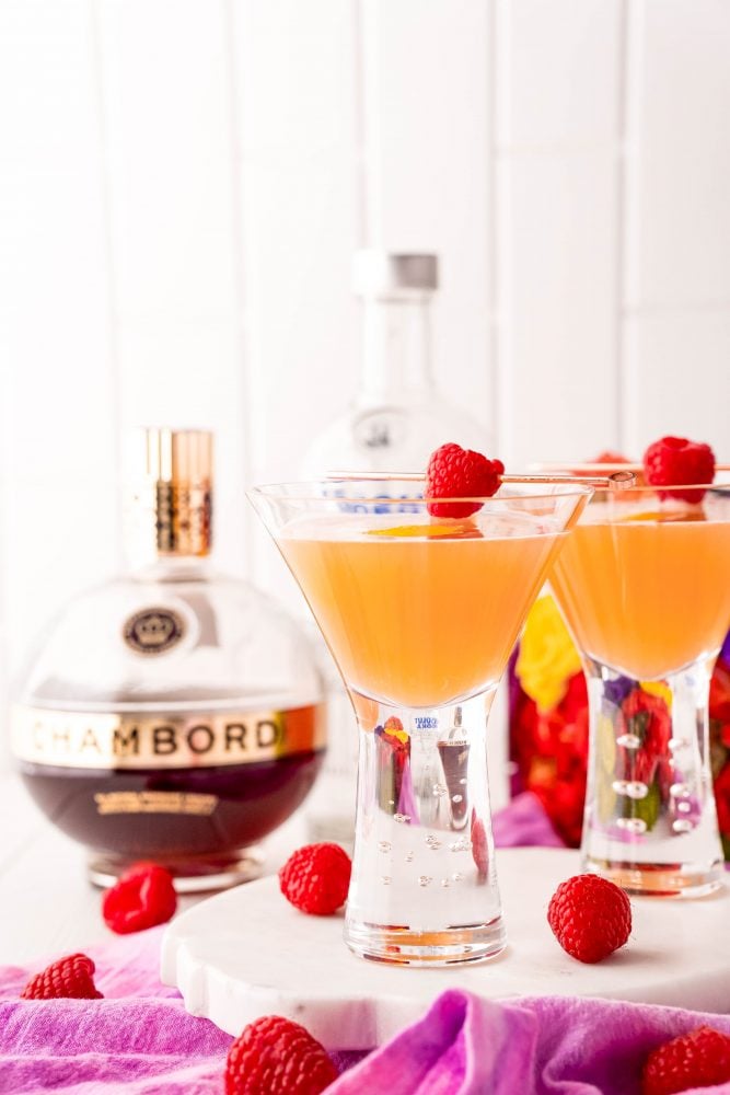 orange martini in a glass with a raspberry stuck across it and chambord in the background.