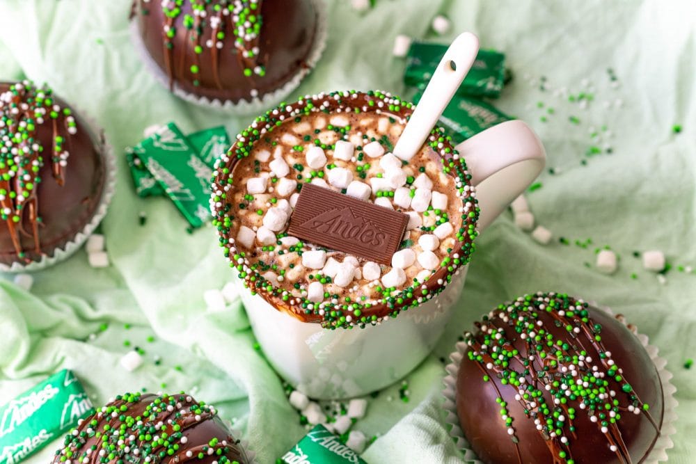 hot chocolate with green sprinkles on the edge with an Andes mint and marshmallows int it and a spoon