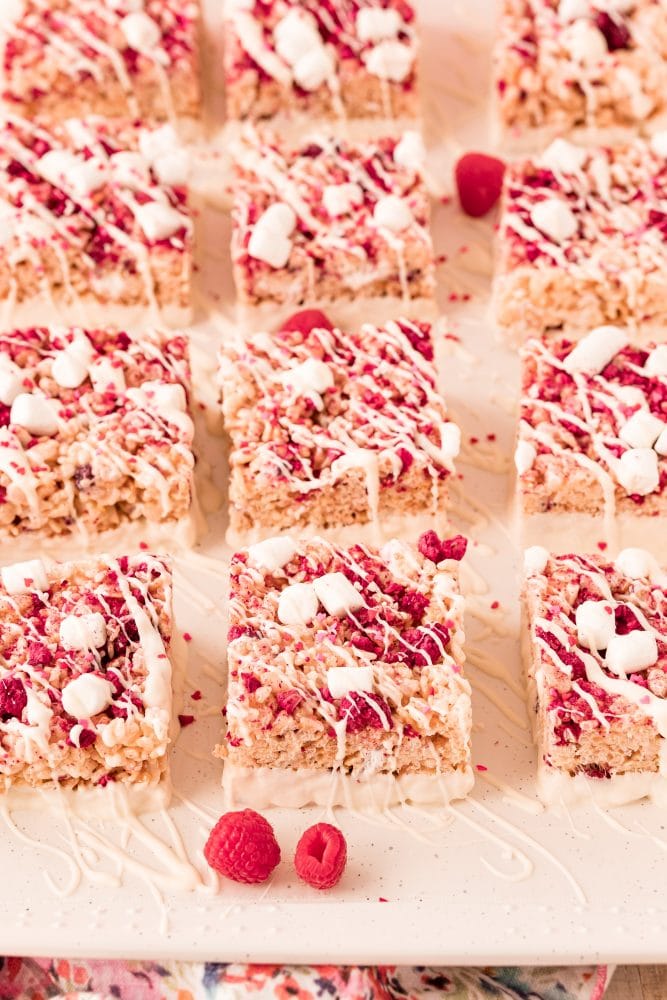 pink rice Krispie treat with marshmallows, freeze dried raspberries in it and white chocolate drizzle set out on a white plate